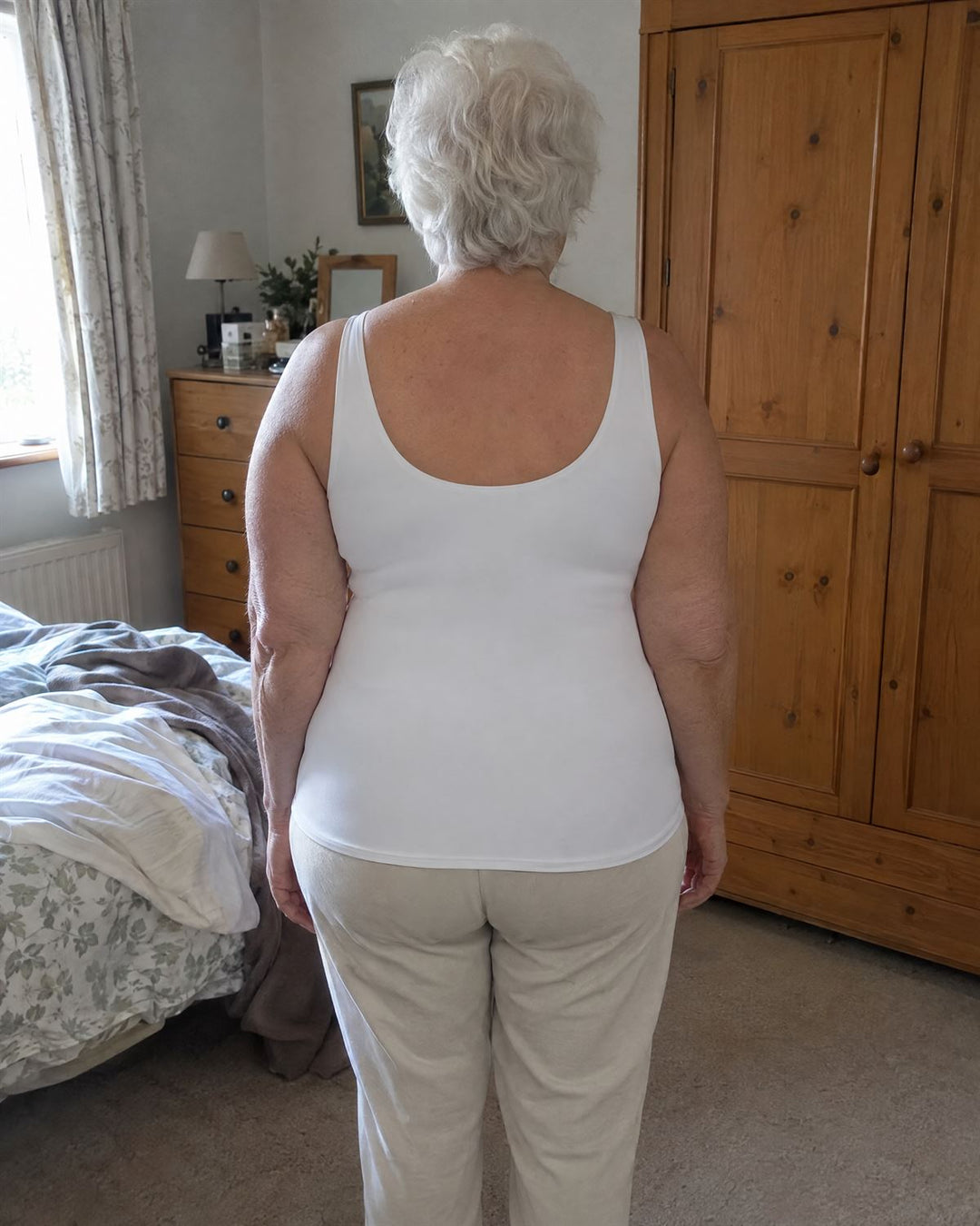 Woman in her seventies in a white SmartFit tank, back view in her bedroom: smooth from shoulders to waist