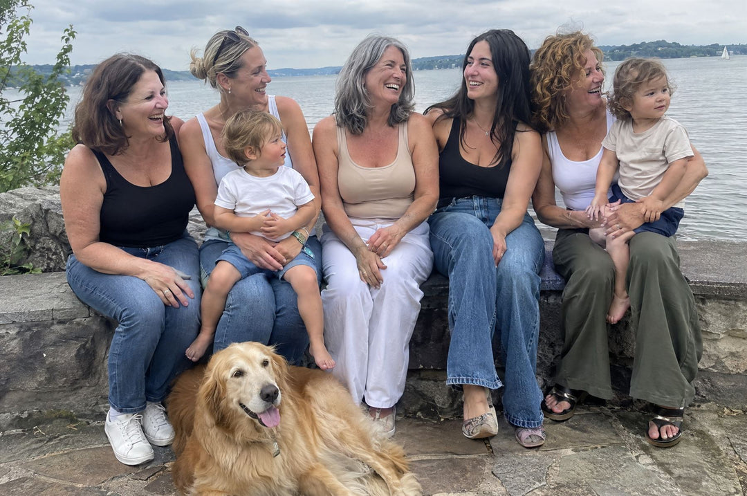 Group of women laughing on a lakeside wall with toddlers and a golden retriever, wearing tanks in black, white and nude